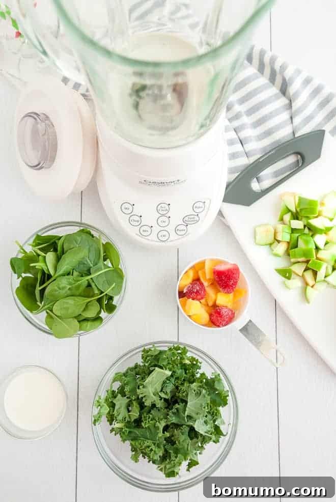 Overhead view of an empty blender, surrounded by bowls of spinach, kale, tropical fruit, and green apples.