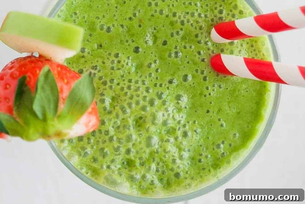Overhead view of a green smoothie in a glass, garnished with a red strawberry and a slice of green apple.