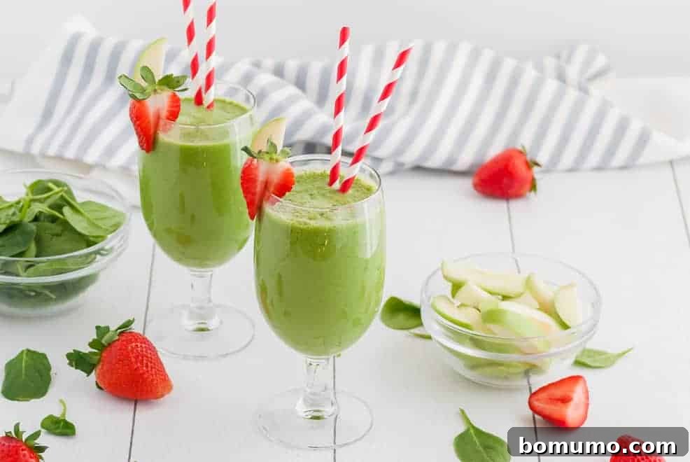 Two green smoothies in stemmed glasses with red and white straws. Also pictured are strawberries, green apples, and a striped linen. 