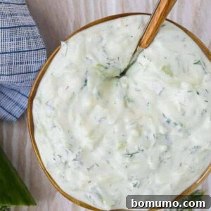 Overhead view of creamy yogurt sauce in a bowl on a wooden background. Cucumbers and dill are placed around the bowl.
