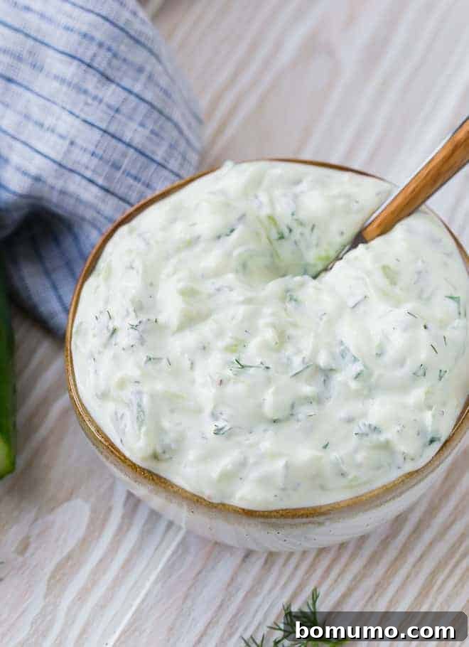 A creamy yogurt sauce made with cucumbers, dill and lemon in a natural colored bowl with a wooden spoon inserted.