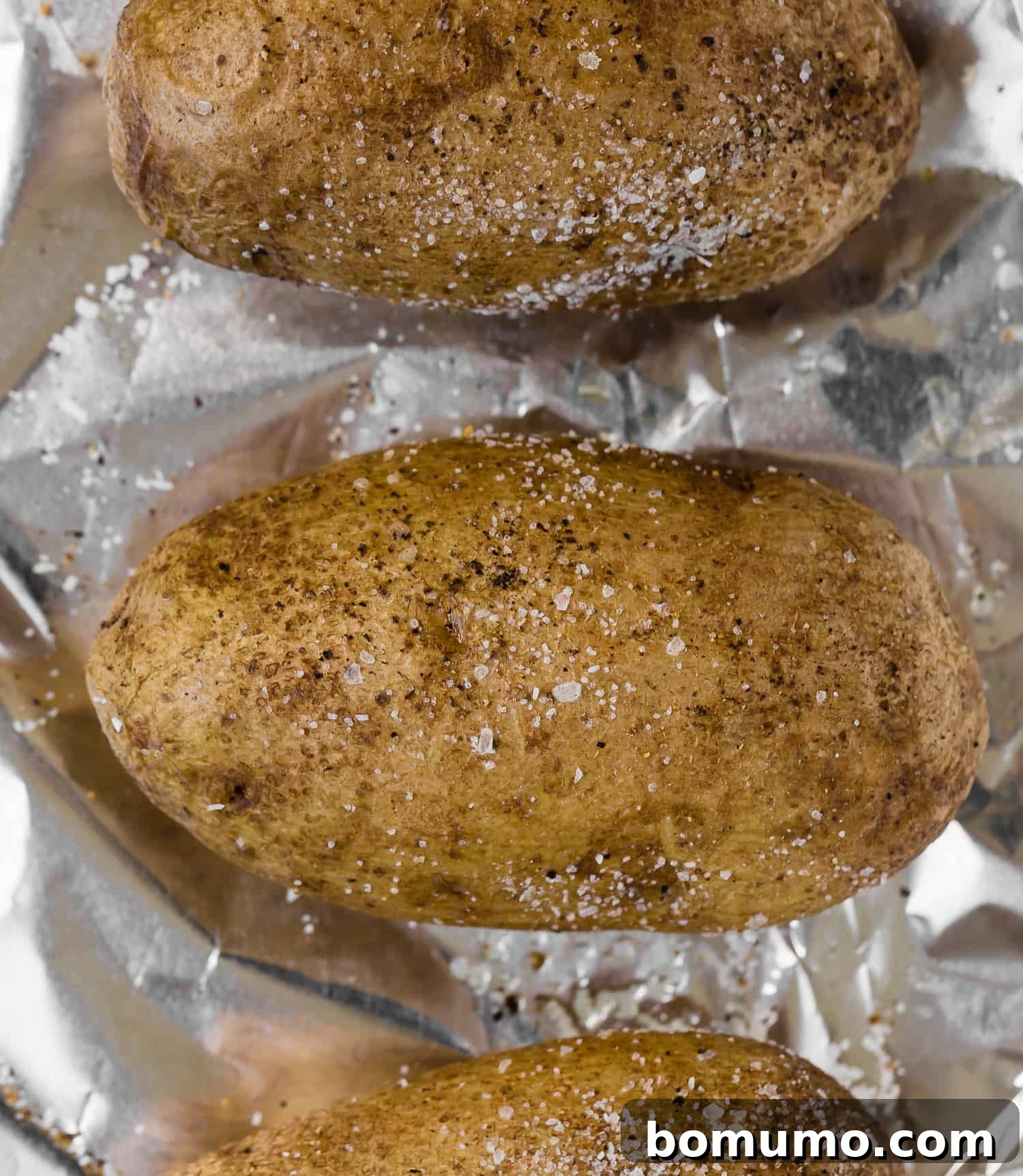 Two golden-brown baked potatoes resting on a simple white plate, seasoned with coarse salt and freshly ground black pepper.