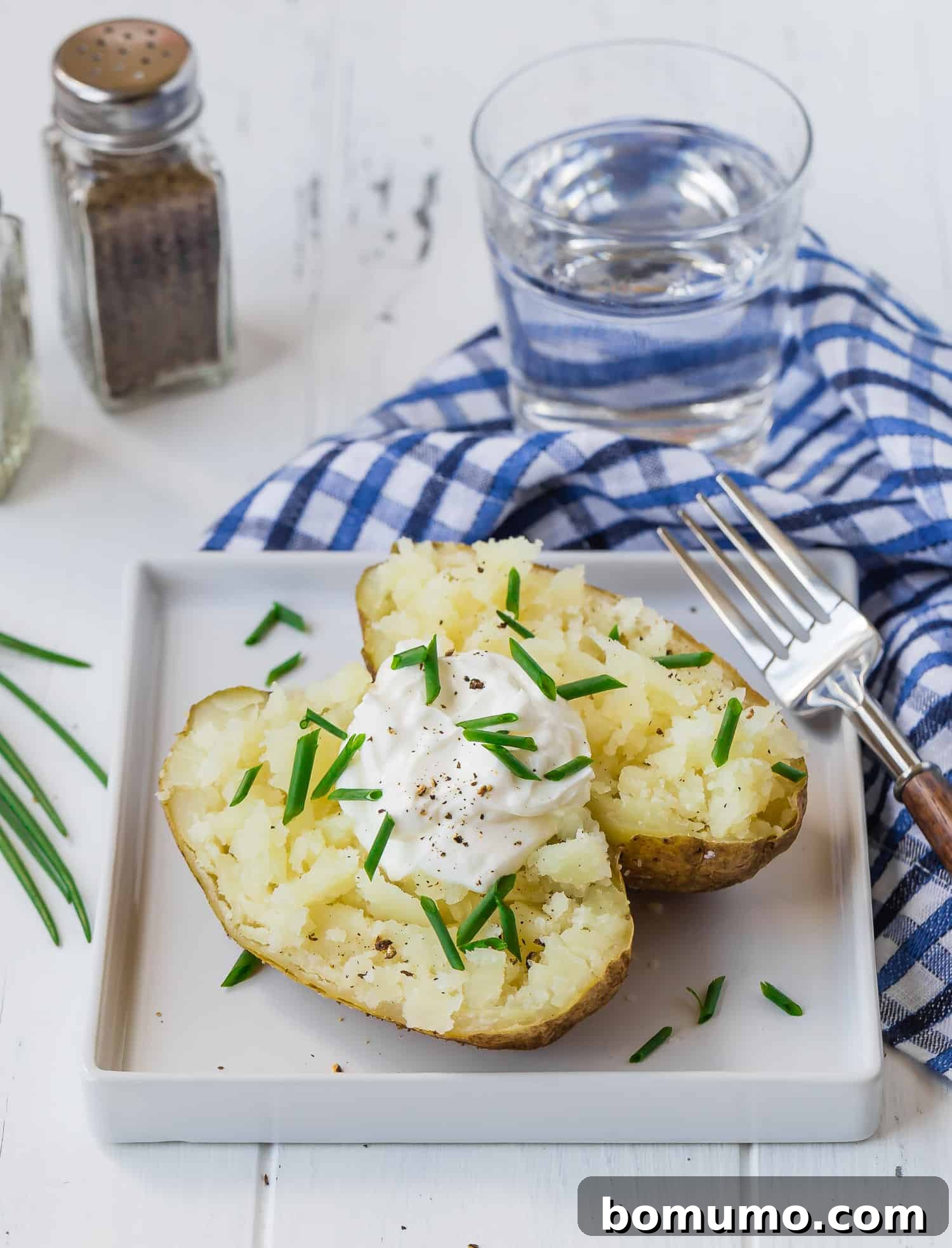 A golden-brown baked potato, perfectly split open, resting on a clean square white plate, awaiting delicious toppings.