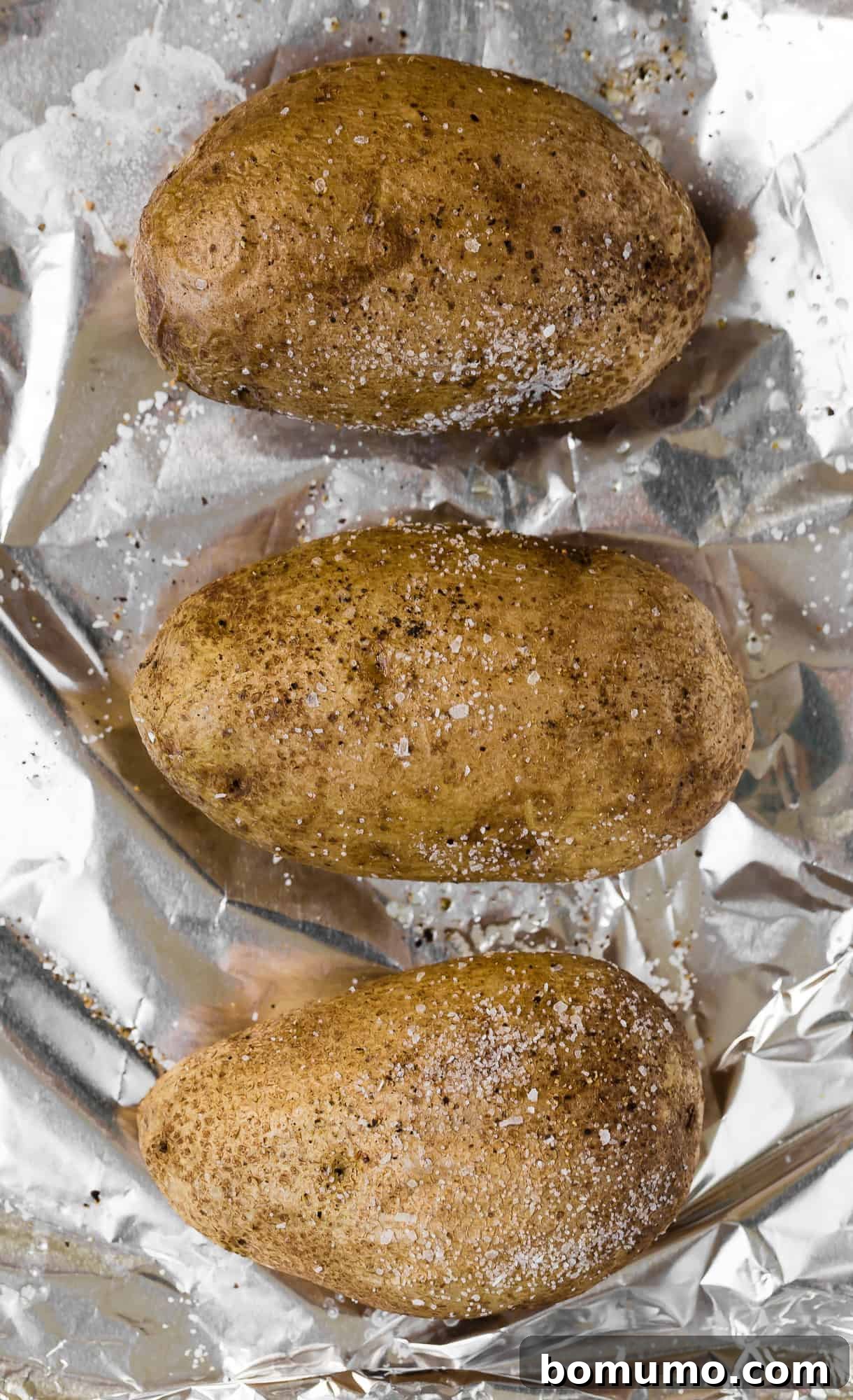 Three perfectly scrubbed and seasoned russet potatoes arranged neatly on a foil-lined baking sheet, ready for the oven.