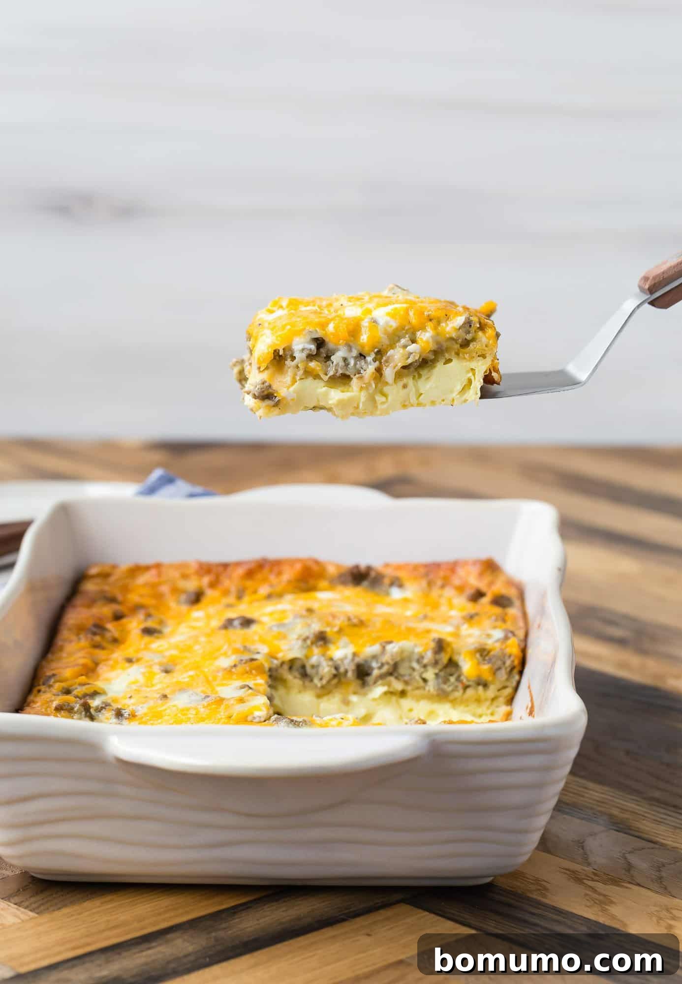 A golden-brown 9x13 pan of crescent roll breakfast casserole, with one slice being lifted on a spatula, showcasing its fluffy layers of egg, sausage, and cheese.