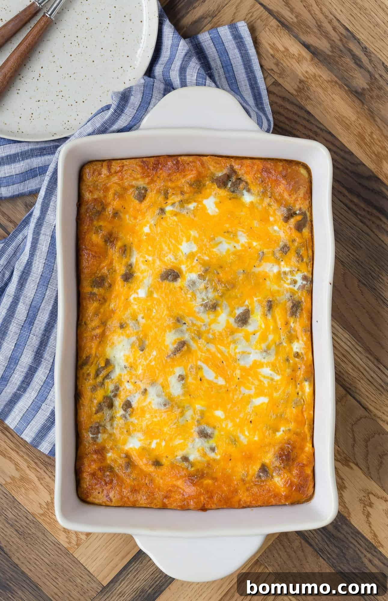 An overhead view of a cheesy crescent roll breakfast casserole in a baking pan, set on a rustic wooden backdrop. Plates, forks, and a blue striped linen hint at a delicious meal awaiting.