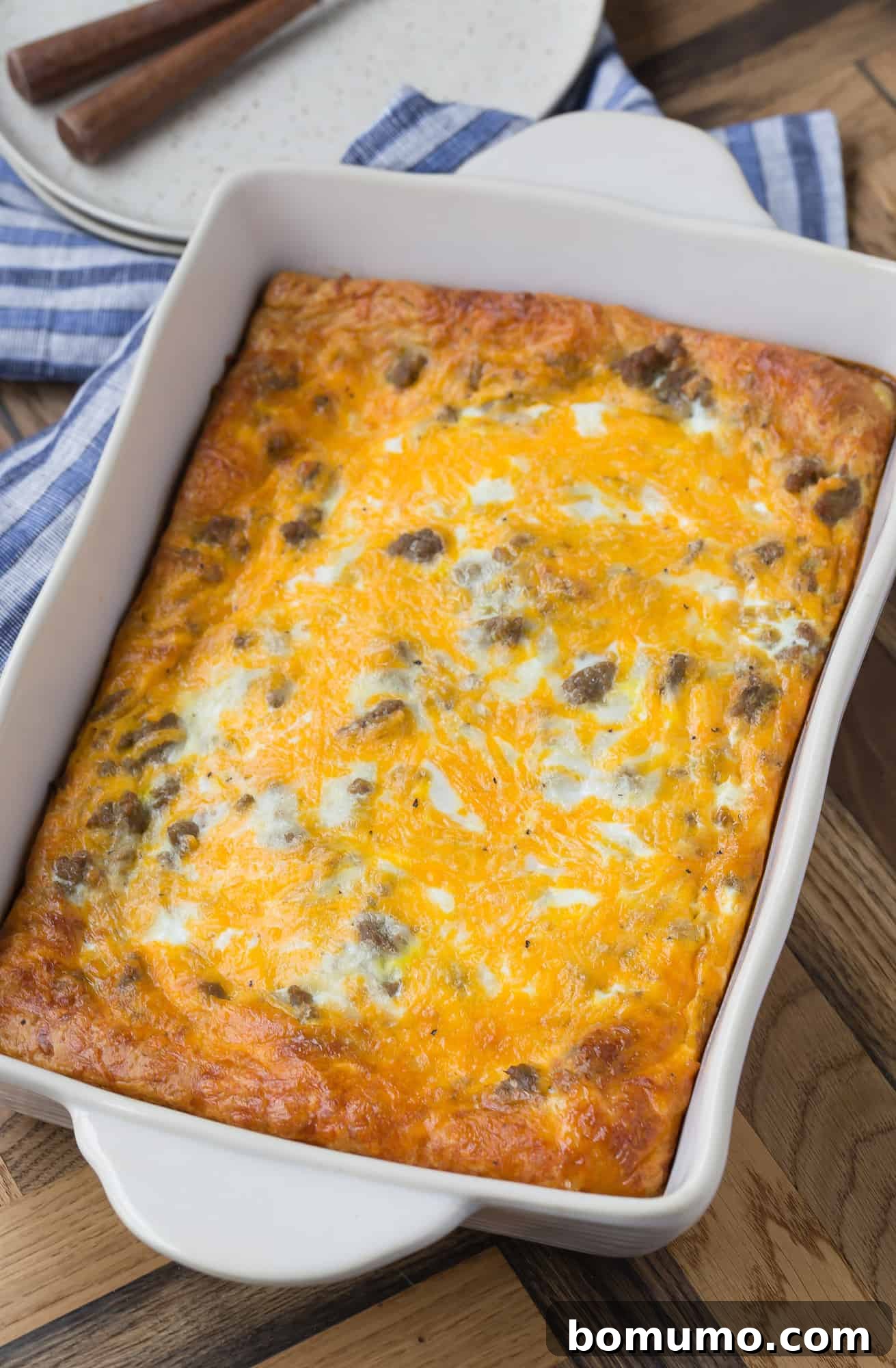 A large white 9x13 baking dish filled with a golden, cheesy sausage and egg breakfast casserole, set against a warm wooden background. Forks and plates are subtly visible in the soft-focus background.