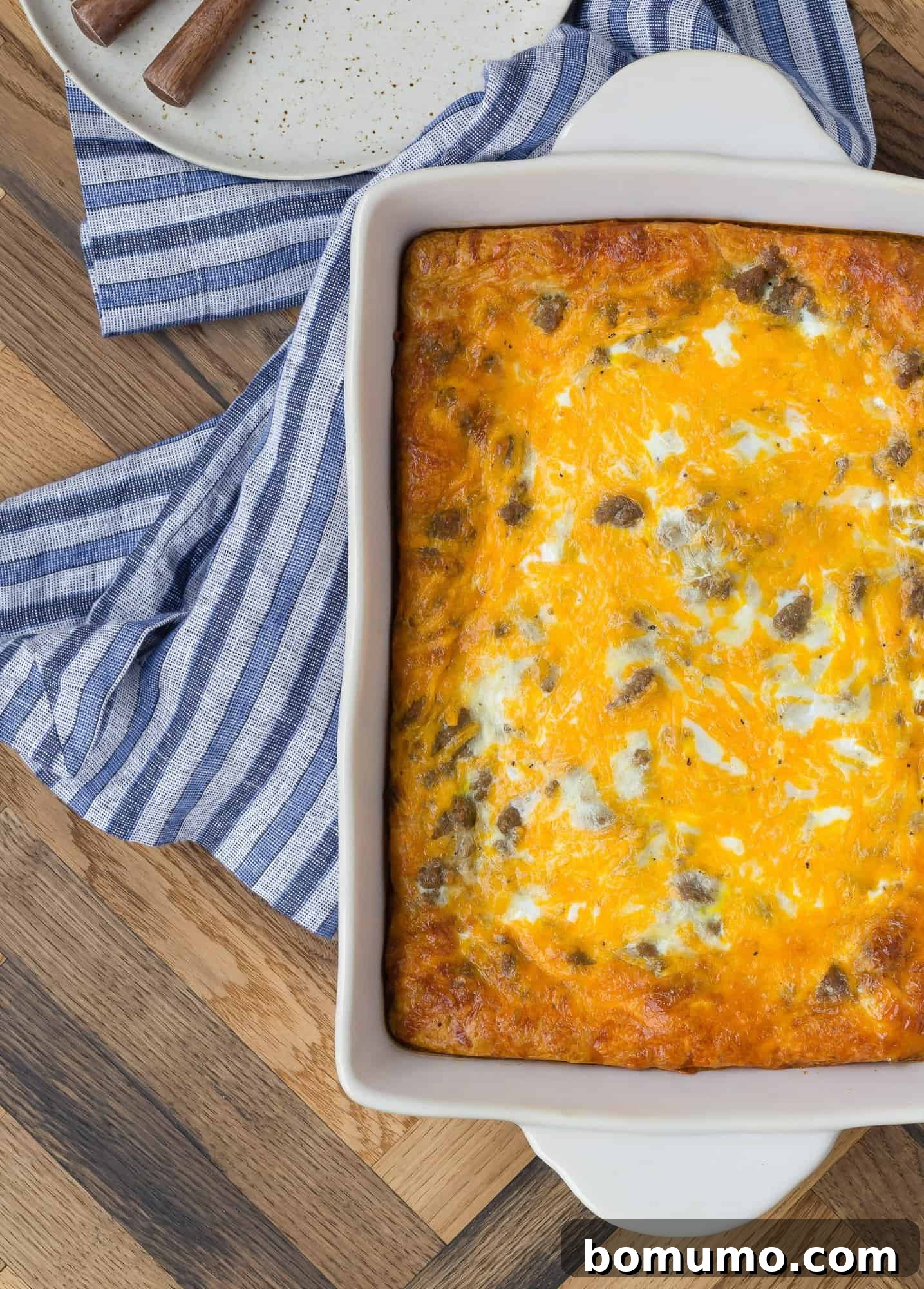 An inviting overhead view of a golden-brown breakfast casserole, presented in its baking pan, resting on a stylish blue striped linen and a warm wooden background.