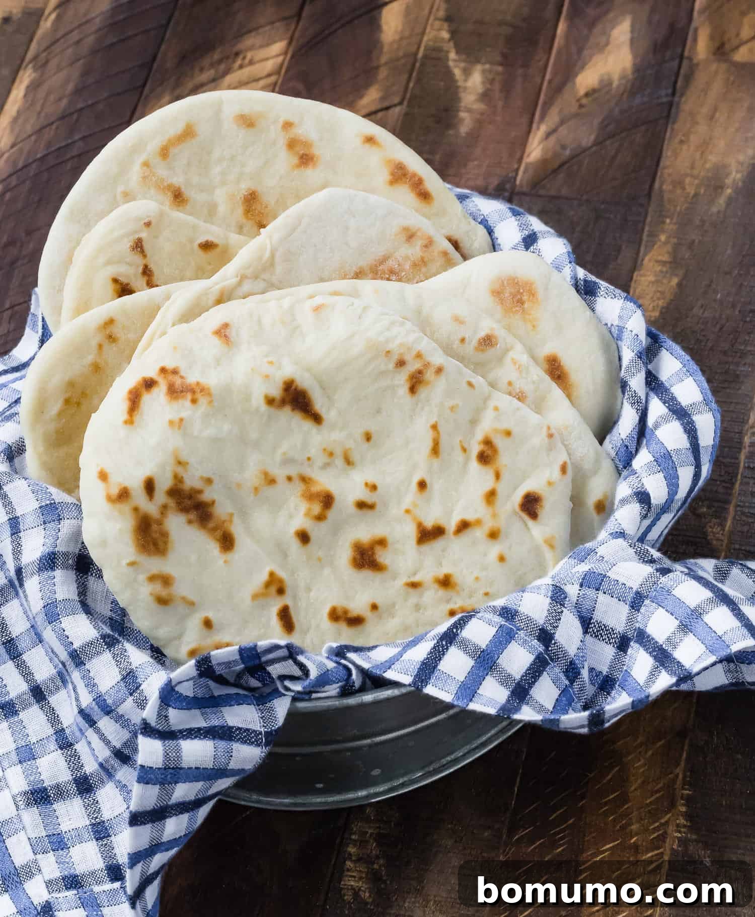 Golden-brown, freshly baked flatbreads neatly stacked inside a decorative tin, ready to be enjoyed. A soft linen peeks out from beneath.