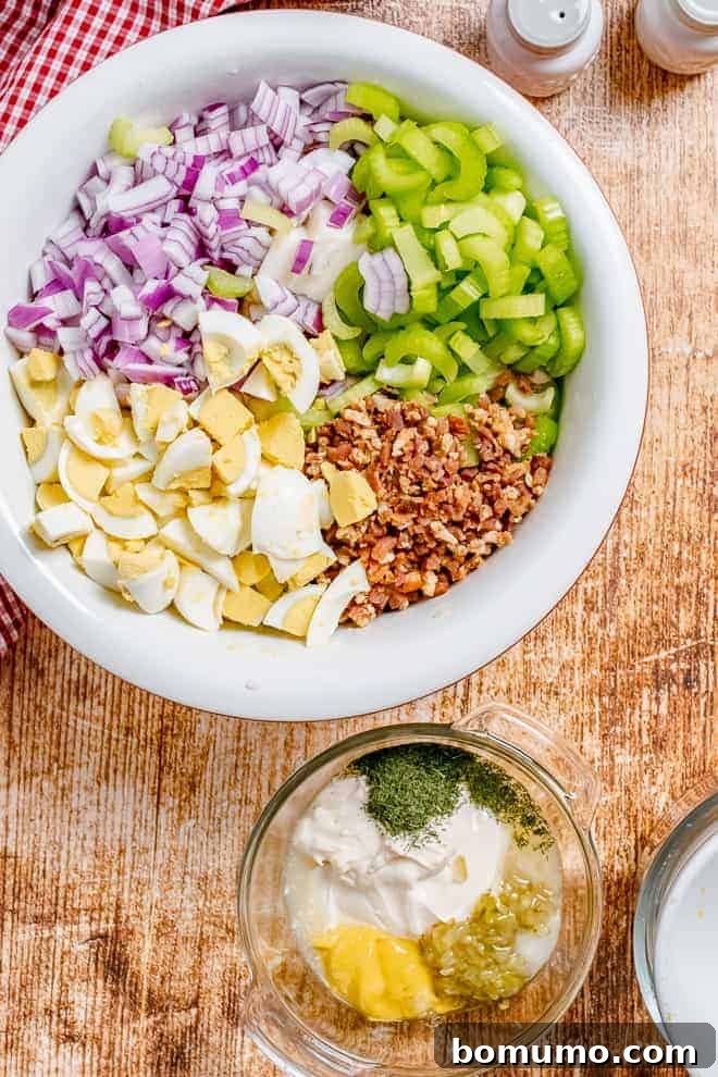 Overhead view of a large bowl filled with chopped boiled eggs, chopped red onion, chopped celery, crumbled bacon. Another small glass bowl sits next to it, filled with a mayonnaise based dressing.