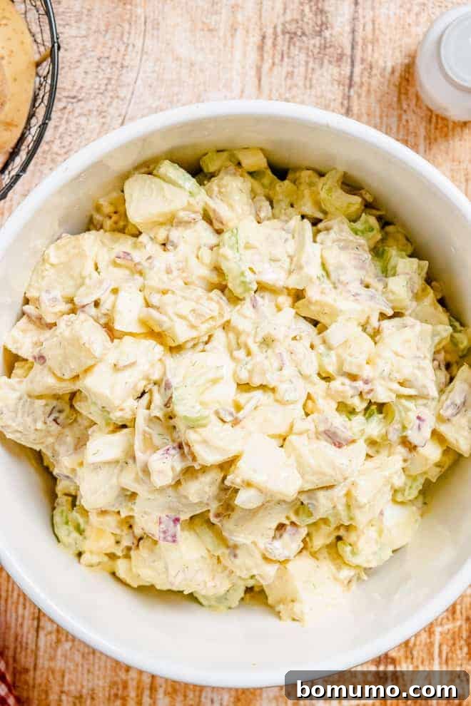 Overhead view of a large white bowl on a wooden background. The bowl is filled with a creamy salad made with potatoes, eggs, celery, bacon, and more.
