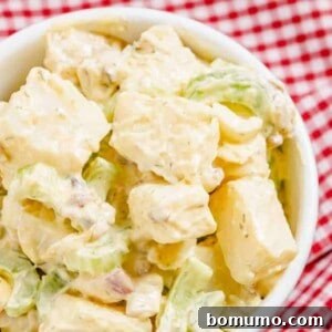 Close up overhead view of a bowl of diced potatoes, celery, onion, bacon, and boiled eggs tossed in a creamy light yellow dressing with flecks of green.