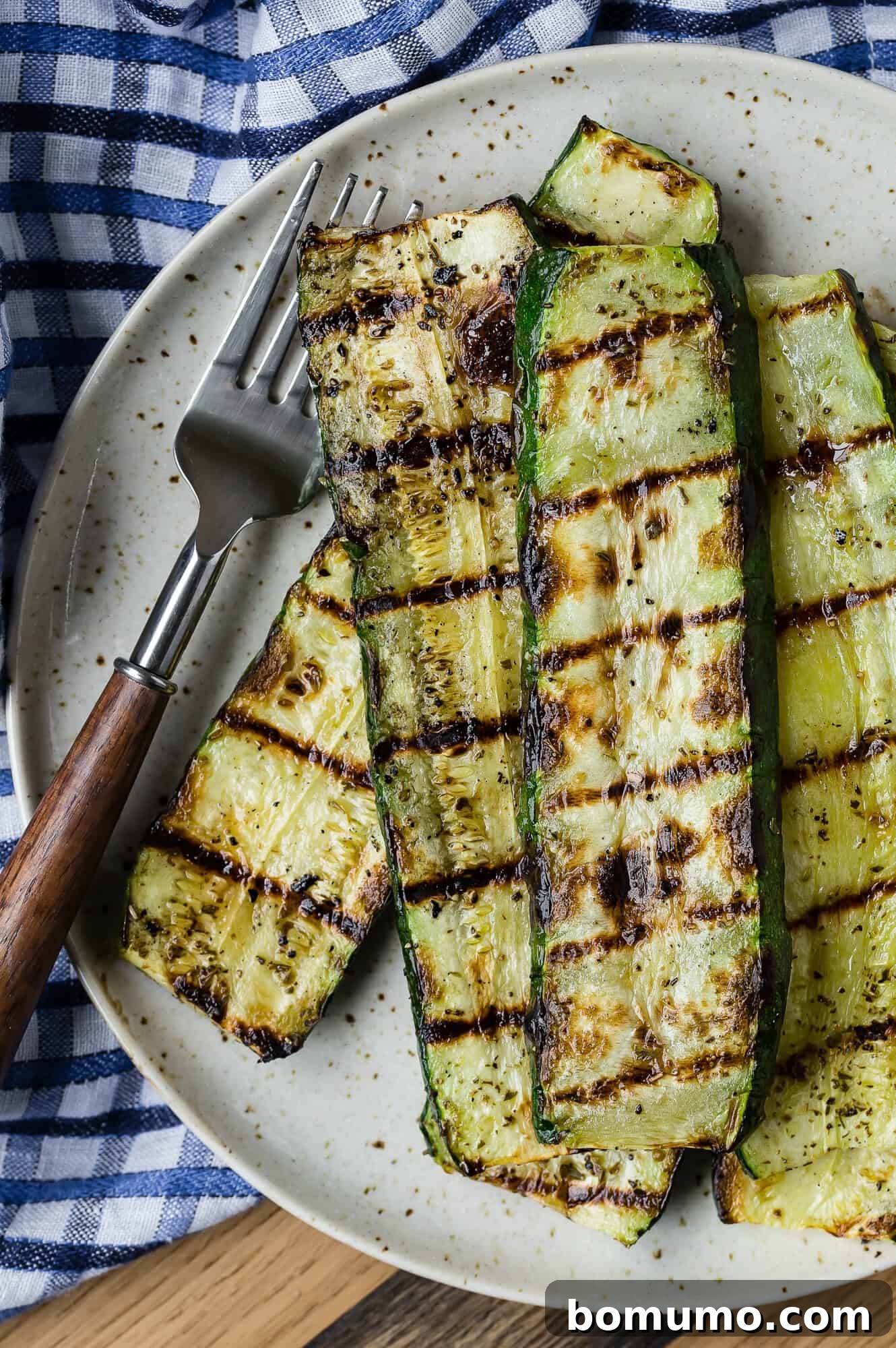 Perfectly grilled zucchini slices, golden-brown and tender, arranged on a plate with a fork, resting on a charming blue checkered tablecloth.