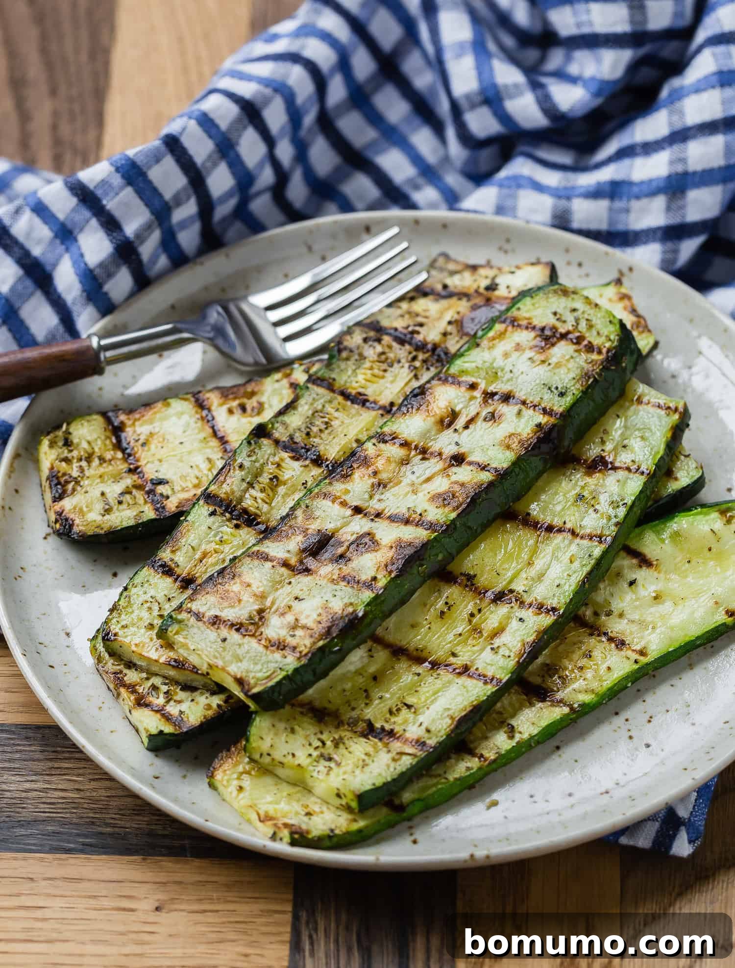 Grilled zucchini slices elegantly presented on a plate next to a fork, with a charming blue checkered cloth creating a rustic backdrop.