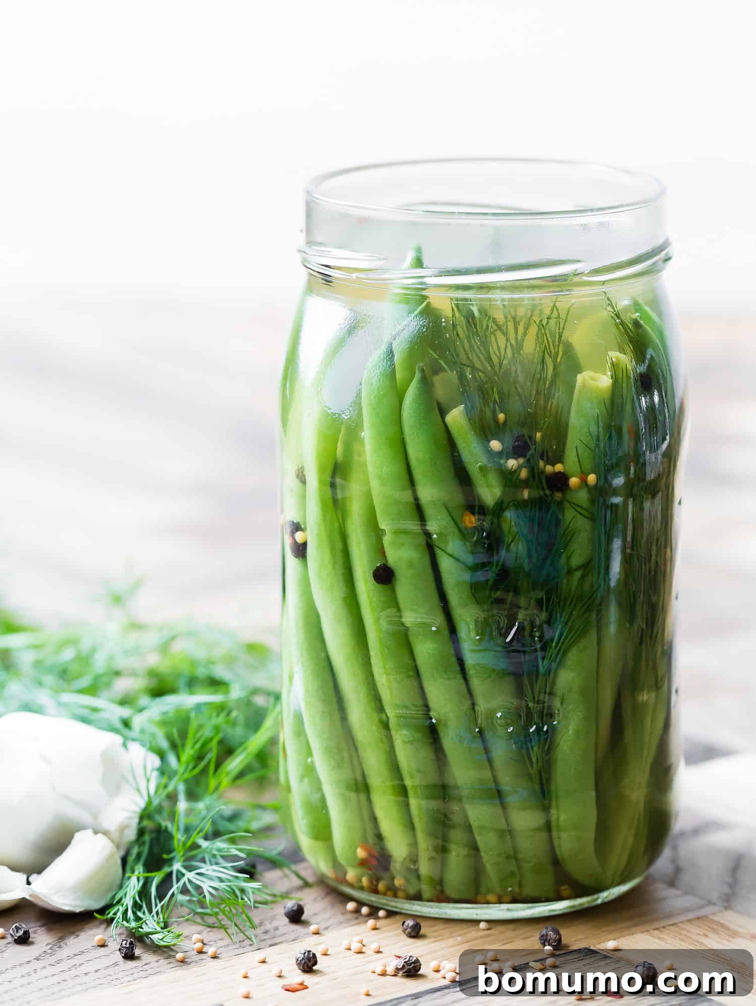 A glass jar filled with vibrant green pickled green beans, fresh dill sprigs, whole garlic cloves, and visible mustard seeds and peppercorns, resting on a rustic wooden surface. The image evokes freshness and homemade goodness.