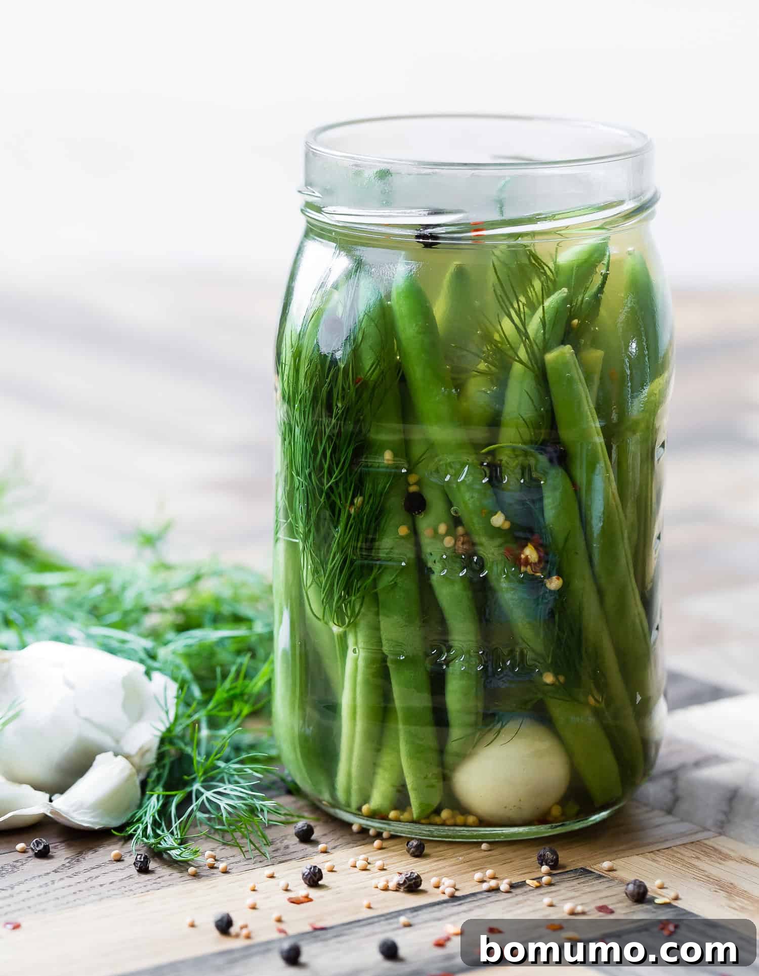 A beautifully arranged glass jar of homemade pickled green beans, garnished with fresh dill sprigs, whole garlic cloves, and visible spices, prominently displayed on a wooden surface. Fresh dill and garlic bulbs are artfully placed beside the jar, emphasizing the fresh ingredients.