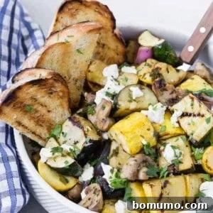 A white bowl filled with grilled zucchini, summer squash, mushrooms, and goat cheese. It is served with grilled bread pieces.