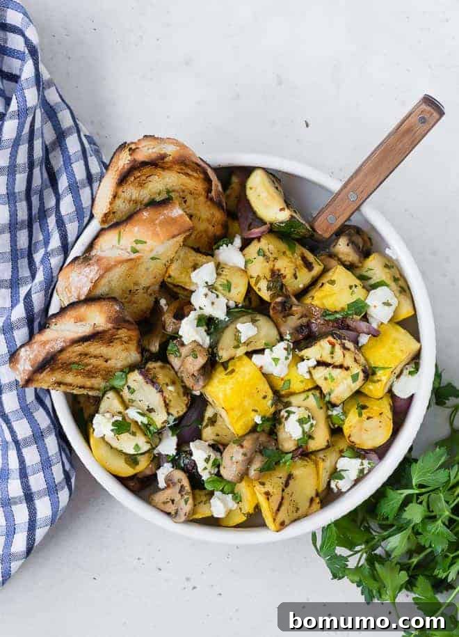 Overhead view of a white bowl on a light grey surface. Bowl is filled with grilled zucchini, summer squash, mushrooms, and goat cheese. It is served with grilled bread. A blue and white linen and fresh parsley is also pictured.