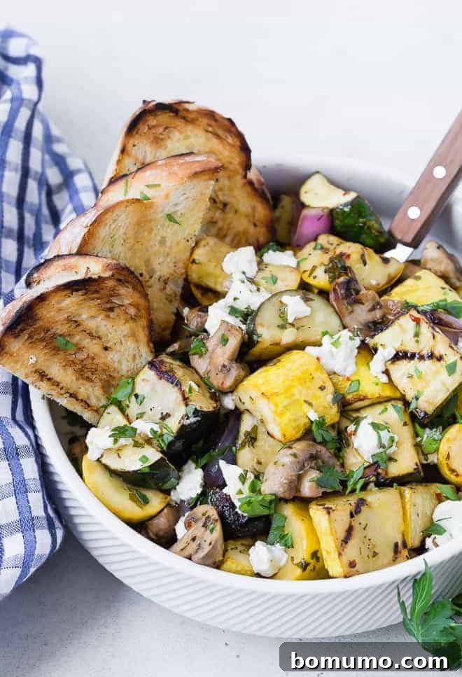 A white bowl filled with grilled zucchini, summer squash, mushrooms, and goat cheese. It is served with grilled bread pieces. 