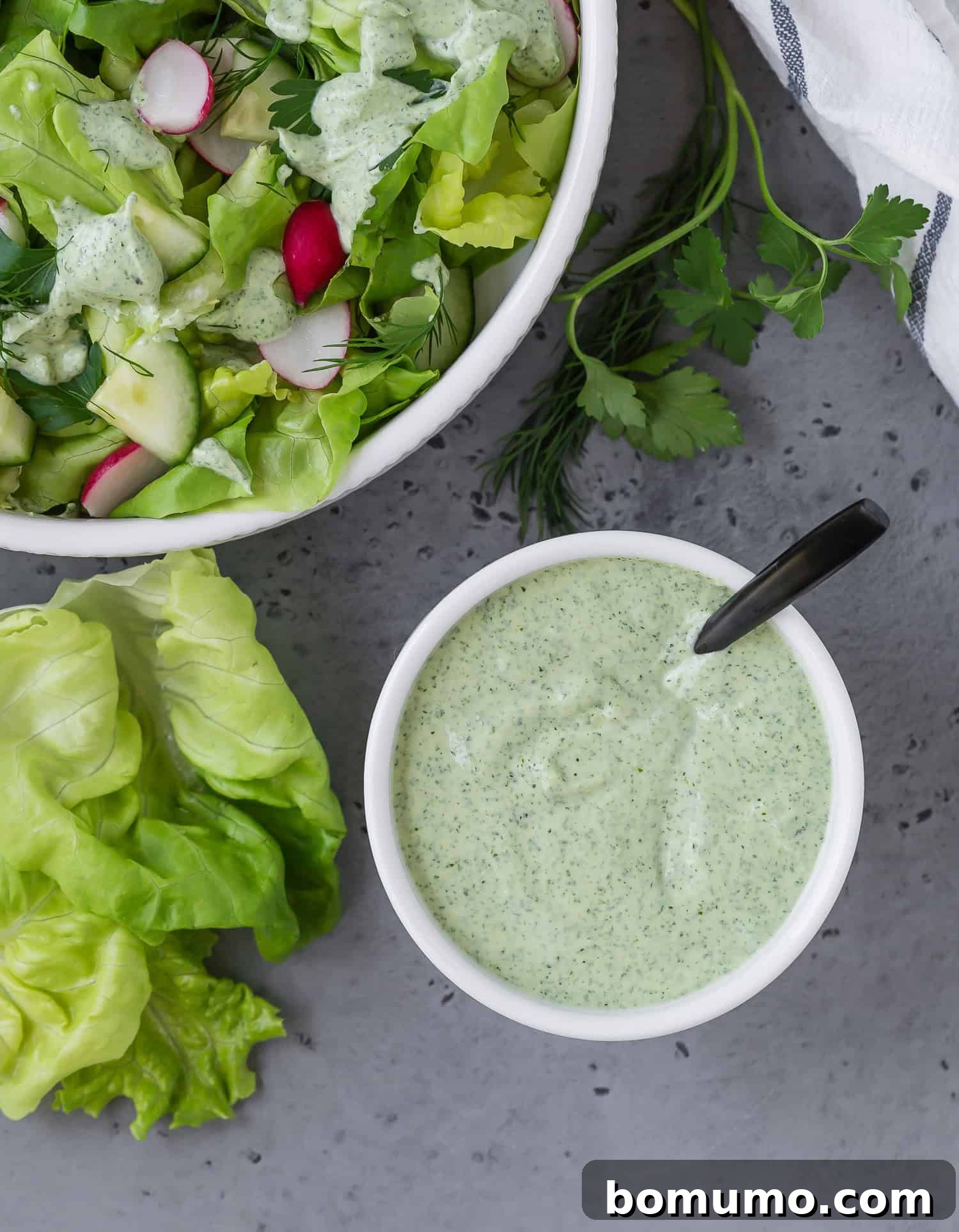 An inviting overhead view of vibrant green goddess dressing in a small white bowl, with a fresh salad subtly blurred in the upper left corner, showcasing its perfect pairing.