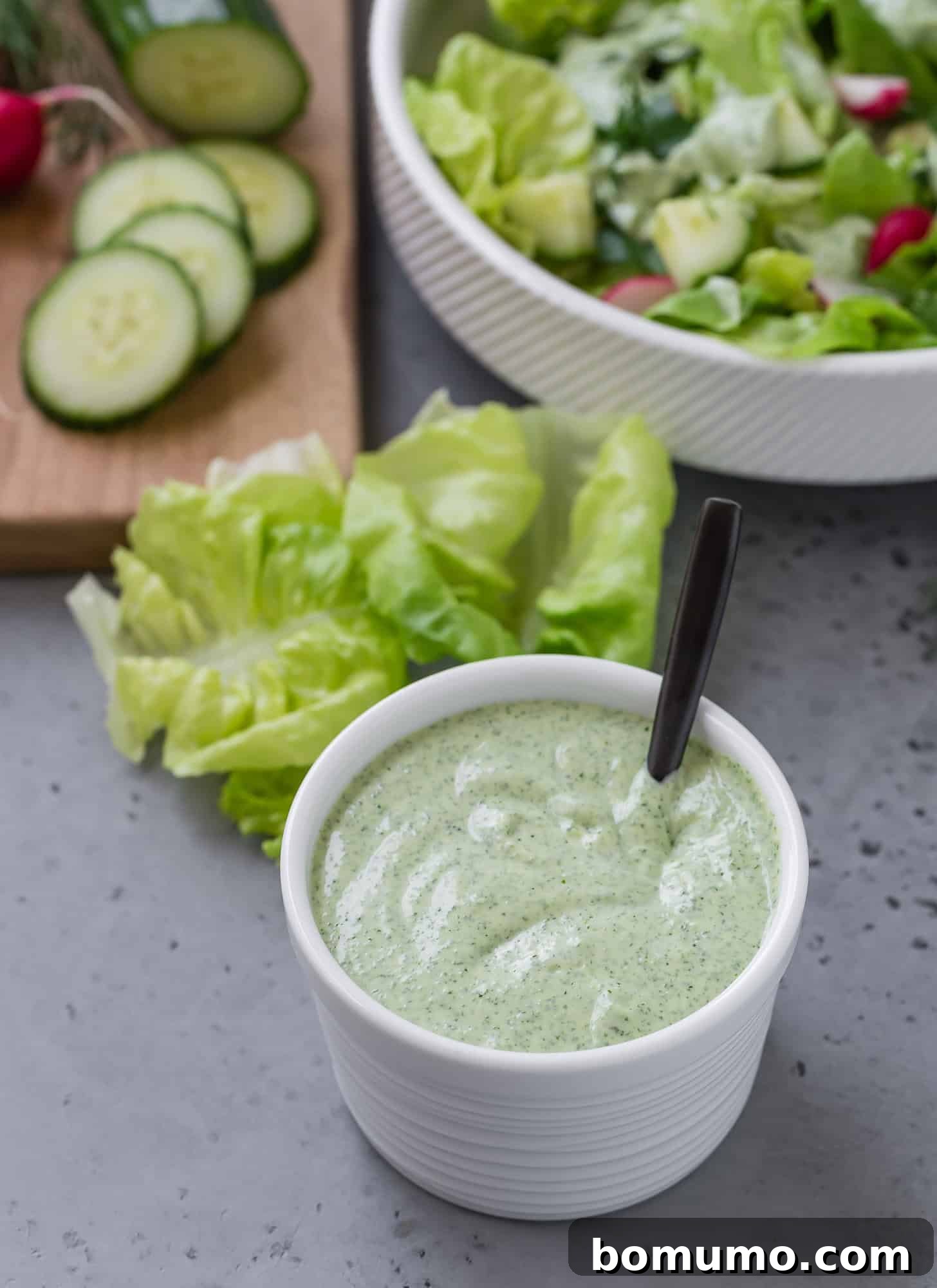 Green goddess dressing displayed in a small white bowl, with a crisp salad featuring cucumbers in the background.