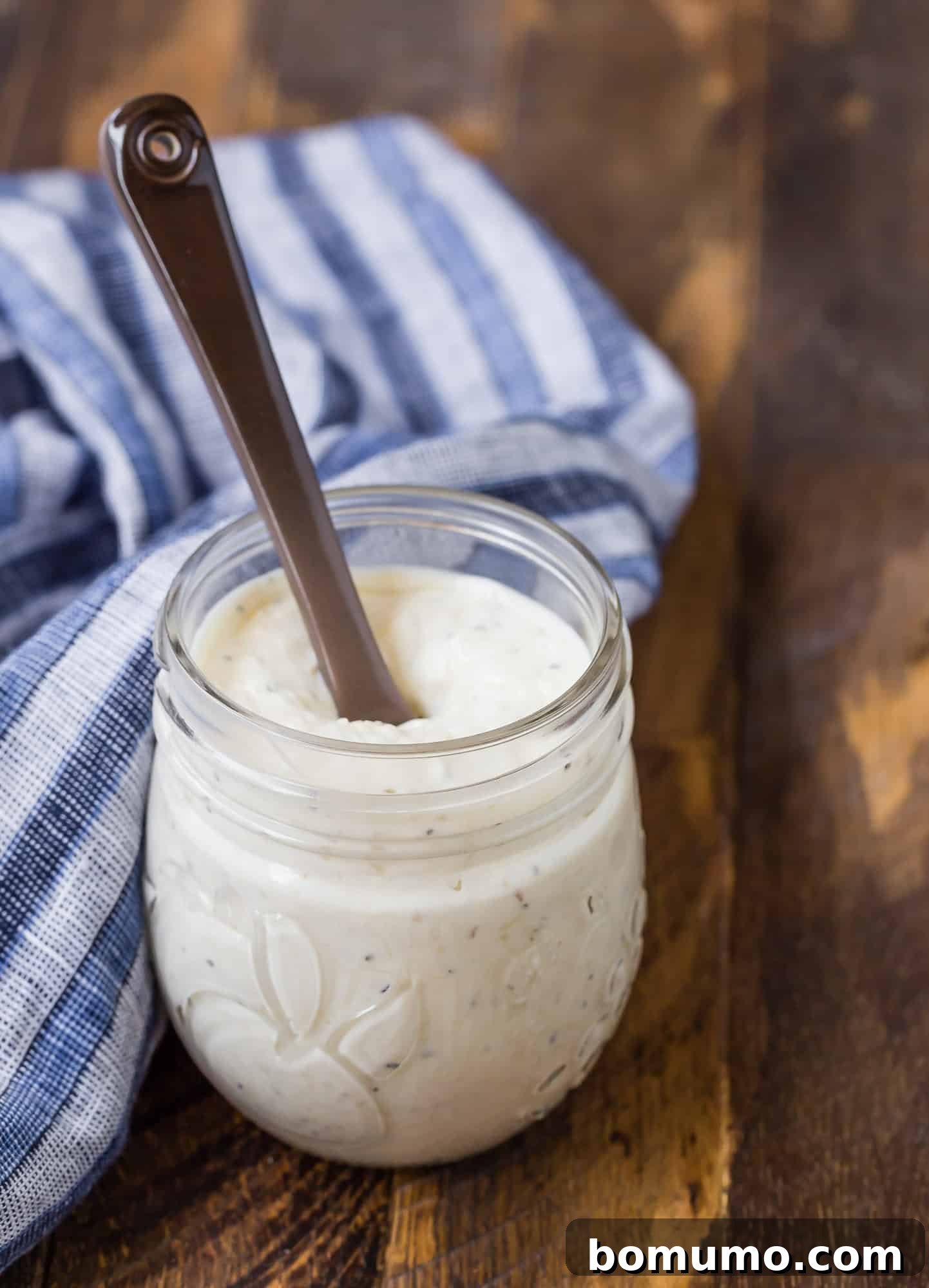 Small glass jar of a creamy caesar dressing on a wooden background. A blue and white linen is also pictured, and a small brown glass spoon. Perfect for your next salad.