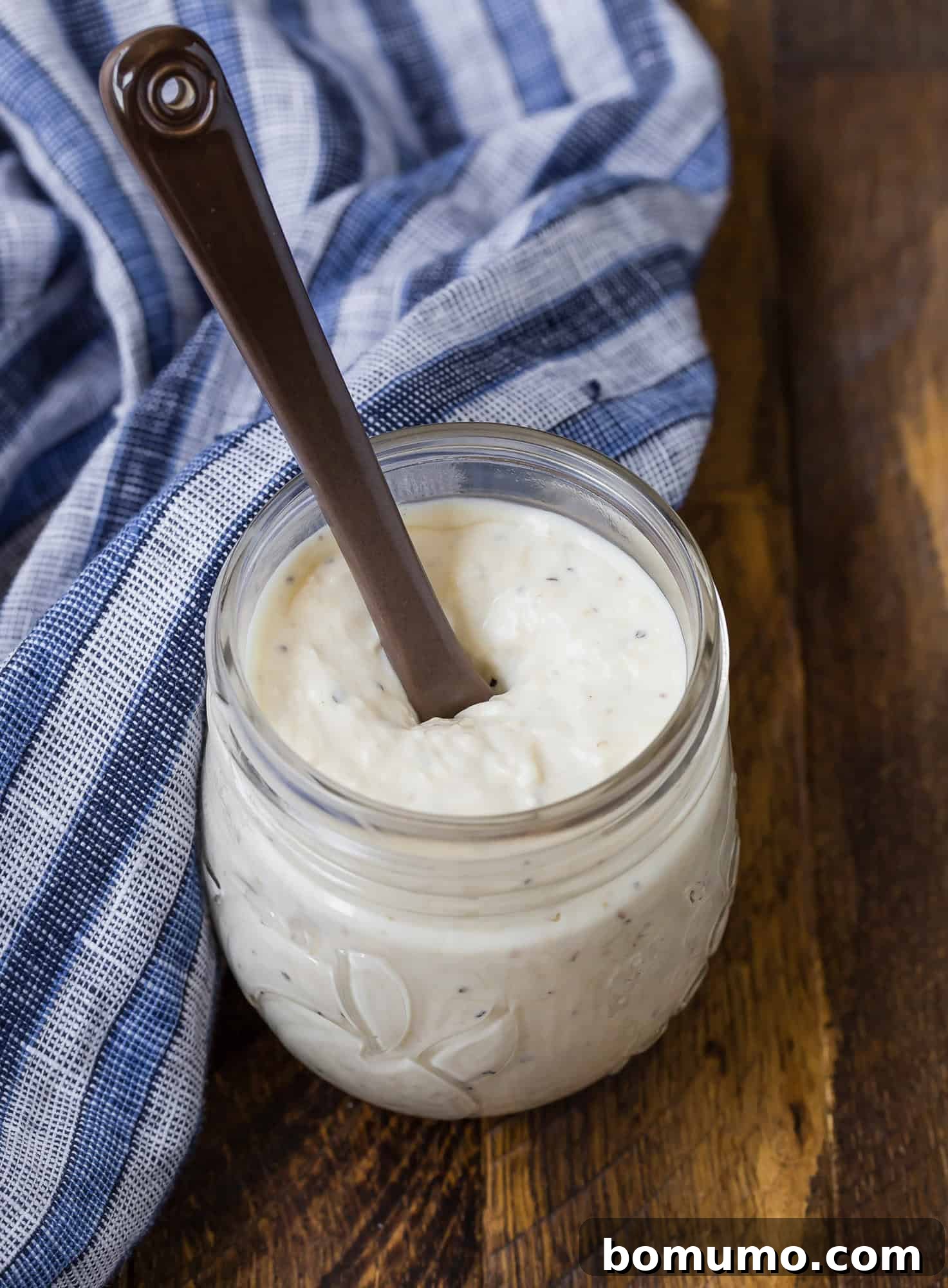 Small glass jar full of creamy dressing. Brown wooden background, blue and white linen. A versatile kitchen staple.