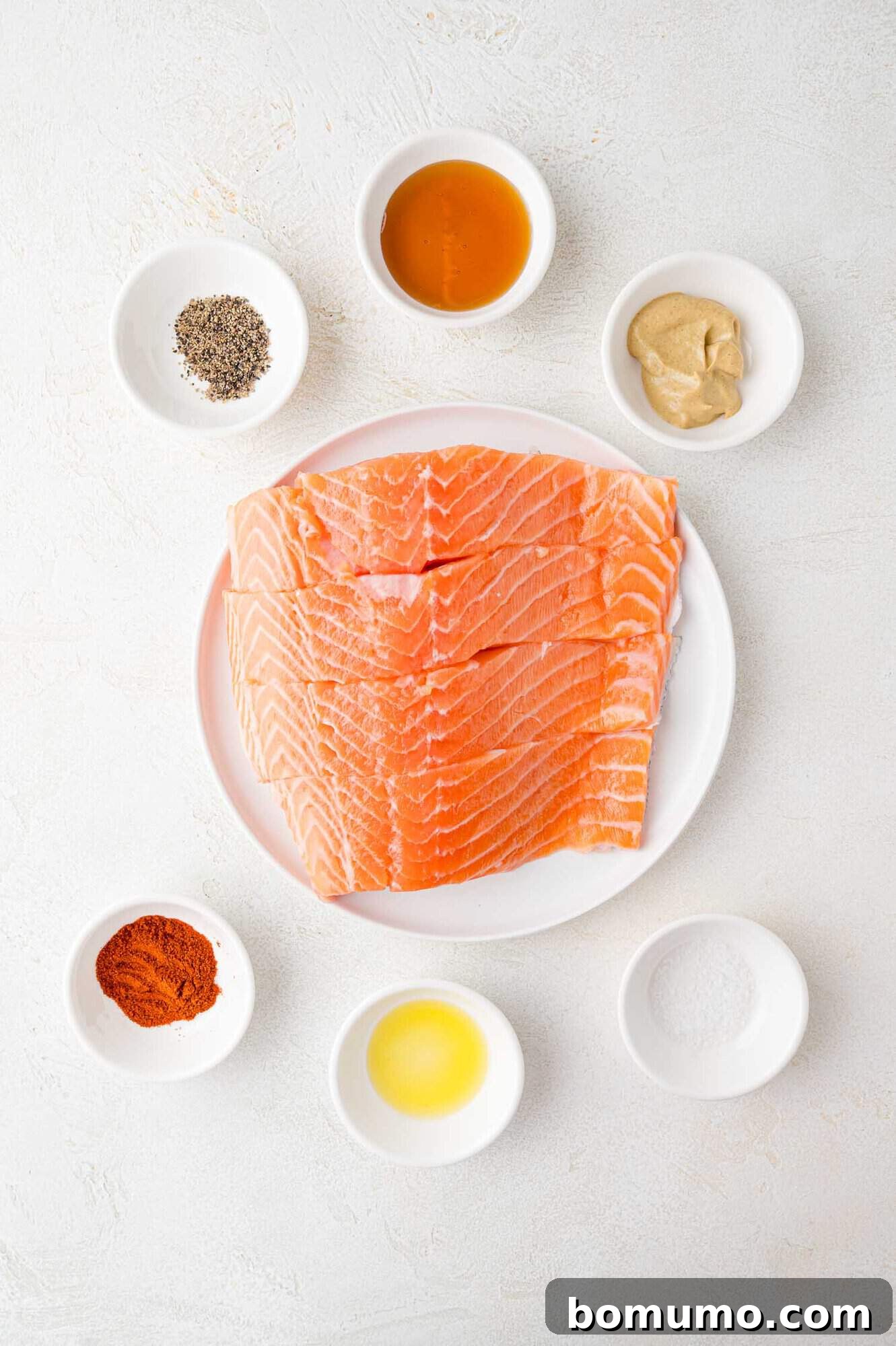 Sliced salmon on a plate surrounded by small bowls containing black pepper, honey, mustard, paprika, and salt.