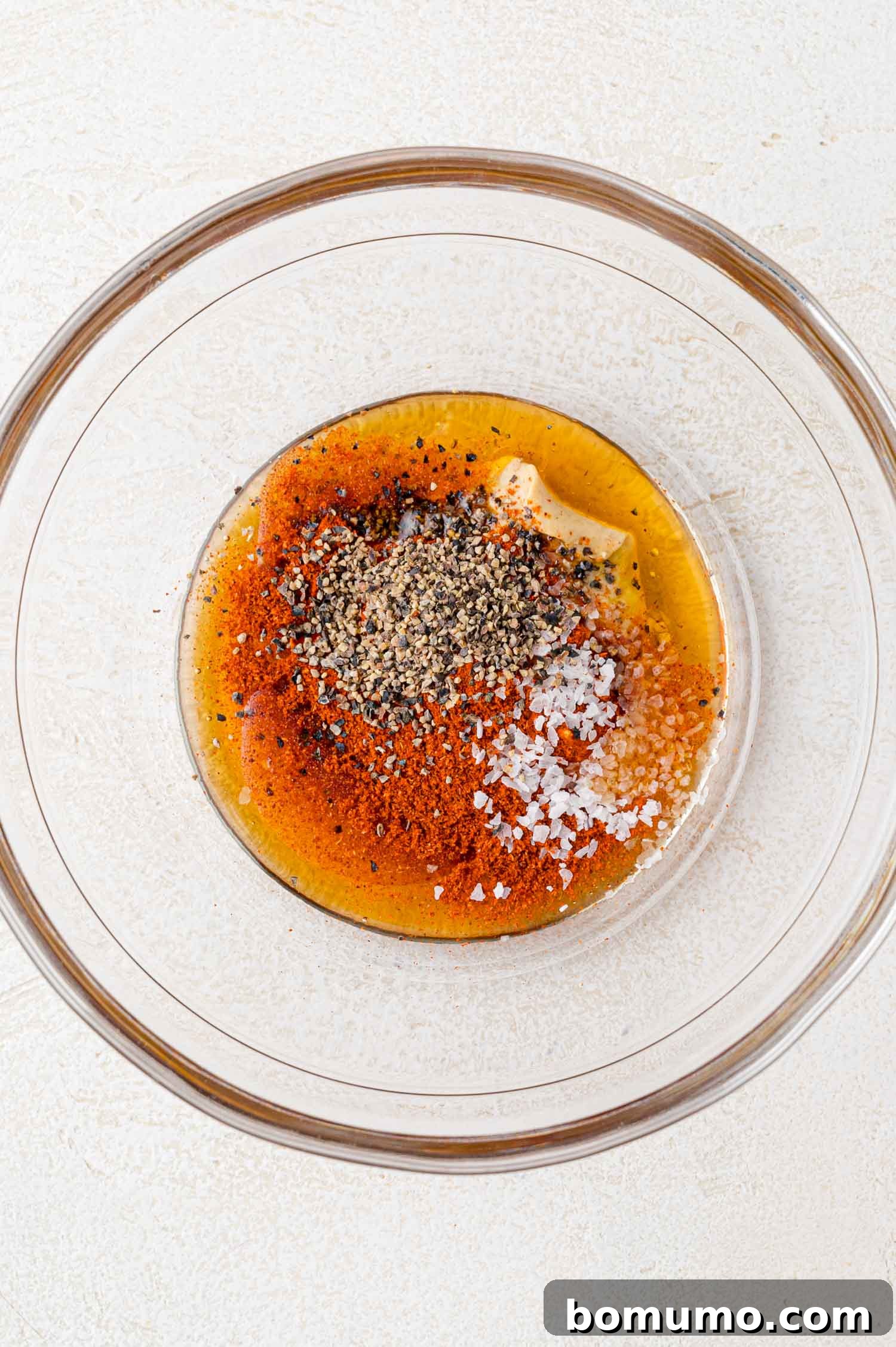 A clear glass bowl containing spices and seasonings, including black pepper, salt, and mustard, on a light textured surface.