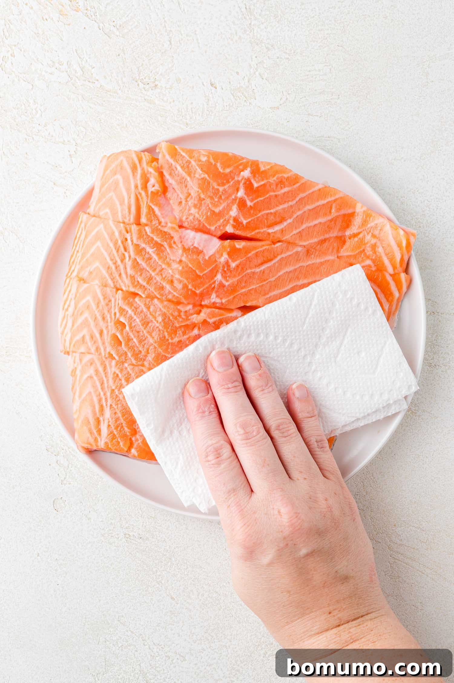 A hand uses a paper towel to pat dry raw salmon fillets on a white plate.