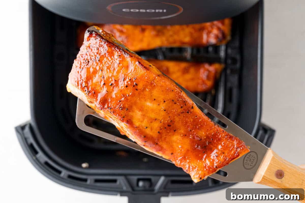 A spatula lifts a glazed salmon fillet from an air fryer basket containing more salmon fillets.
