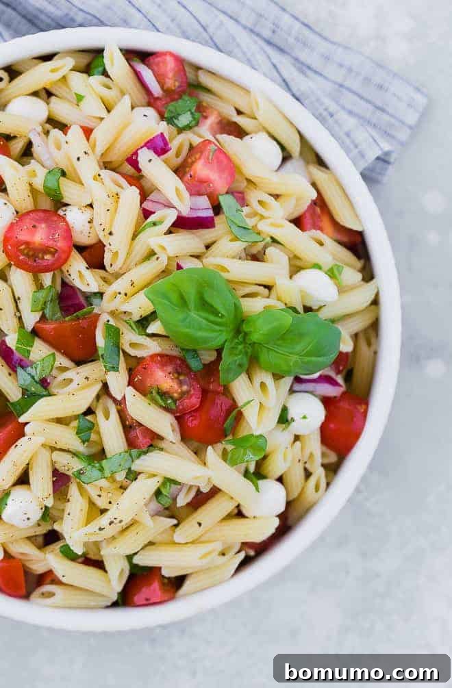 Close up partial view of a bowl of pasta salad with tomatoes, fresh mozzarella, red onion, and fresh basil.