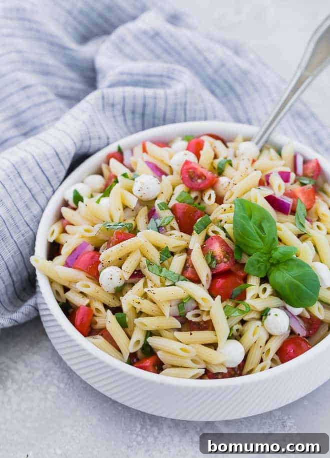A white bowl full of mini penne pasta, grape tomatoes, pearl mozzarella, and fresh basil. A large silver serving spoon is also in the bowl.