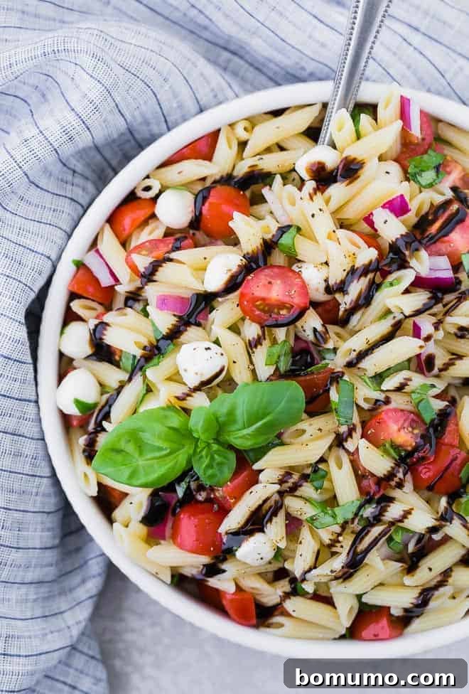 A large white bowl full of caprese pasta salad with tomatoes, mozzarella, red onions, and fresh basil. The salad is drizzled with balsamic reduction.