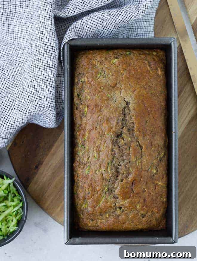 Overhead view of a golden brown loaf of zucchini bread in a metal loaf pan, next to a small bowl of freshly shredded zucchini.