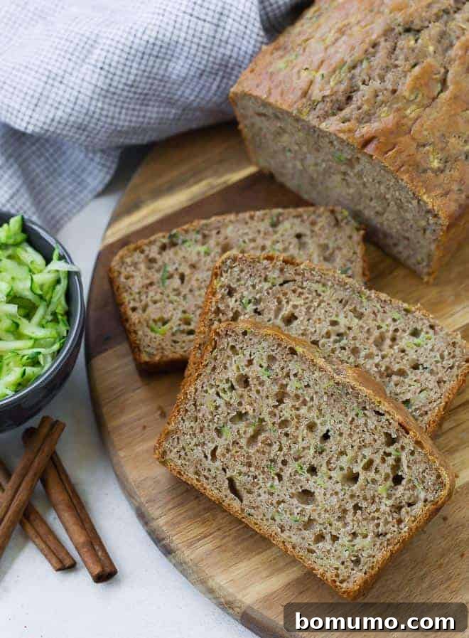 Sliced zucchini bread on a wooden surface, accompanied by a small bowl of shredded zucchini and three cinnamon sticks.