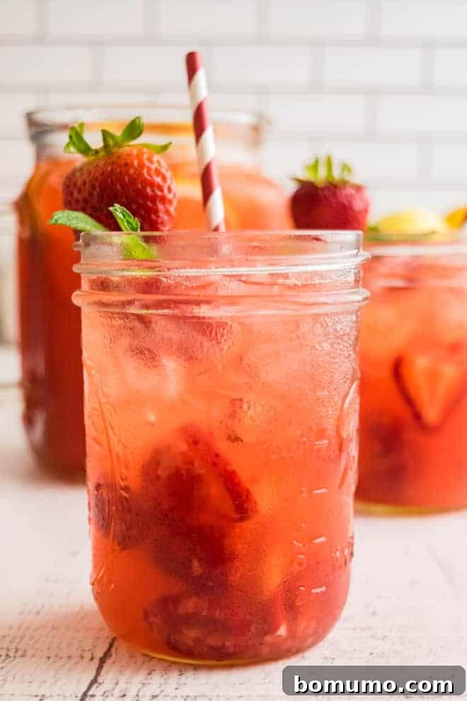 A light red drink on ice in a small mason jar. It is garnished with a fresh strawberry, mint leaves, and a red and white striped paper straw.