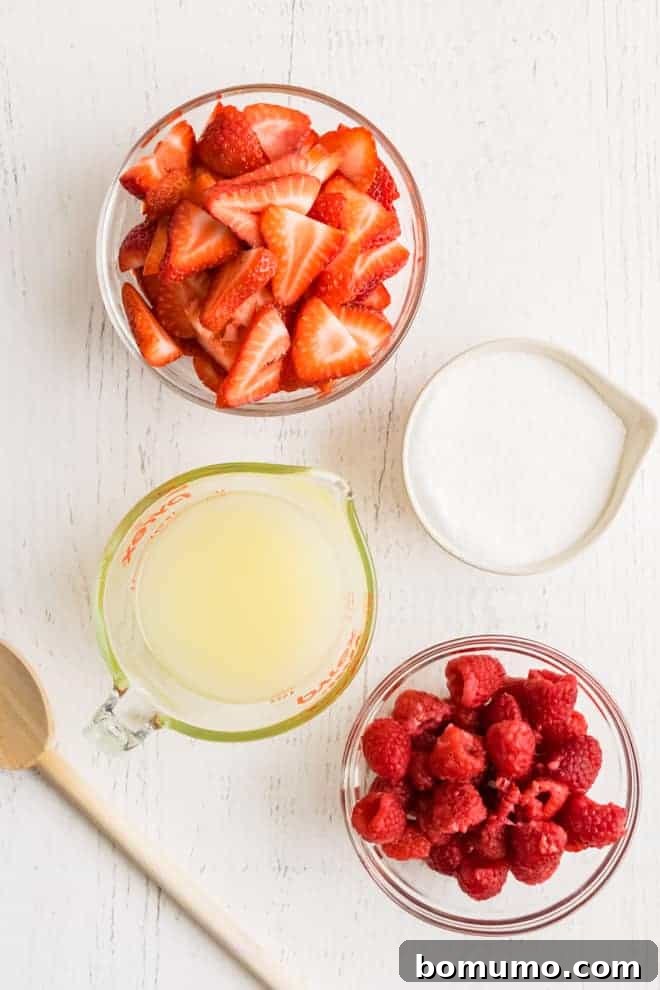 Overhead image of four glass containers, one with strawberries, one with sugar, one with lemon juice, and one with fresh raspberries.