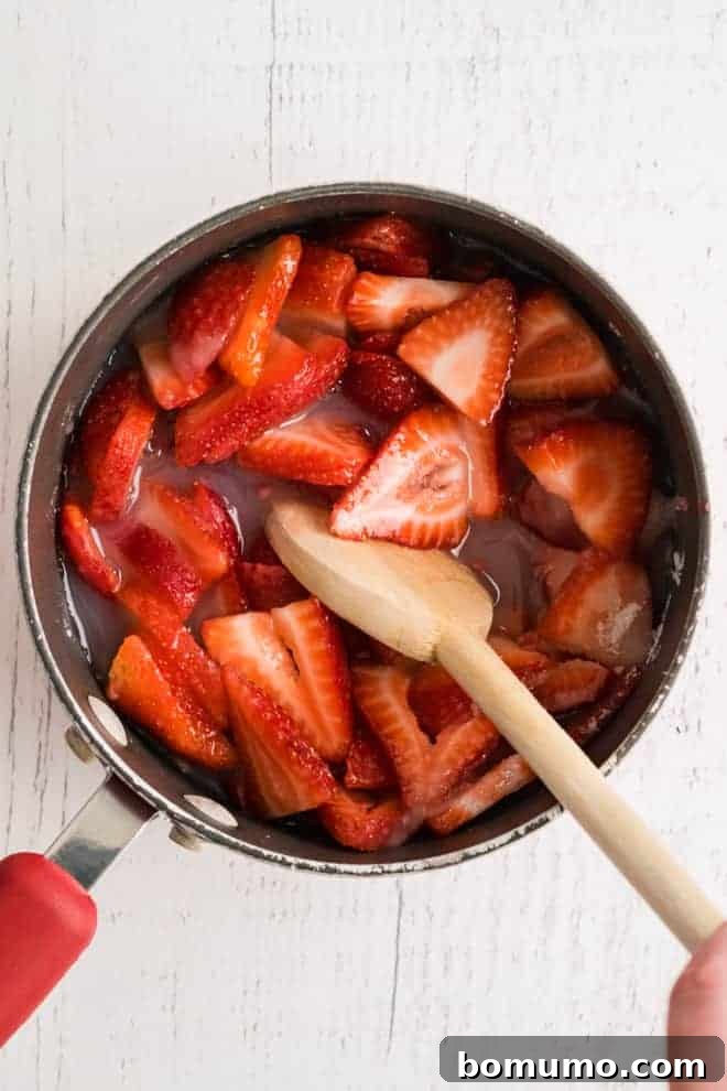 Overhead view of a sauce pan full of strawberries and red liquid, being stirred with a wooden spoon.