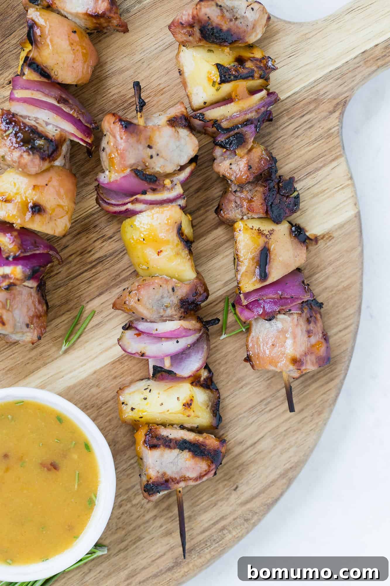 Platter of grilled pork kebobs with apples and onions, garnished with fresh herbs and a side of apricot dipping sauce.
