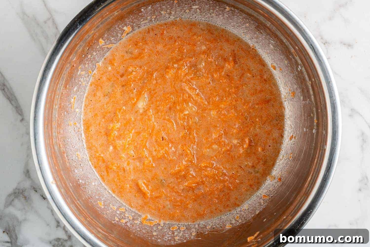 A metal mixing bowl containing the wet ingredients for carrot raisin muffins, including mashed banana, milk, oil, egg, and grated carrots.