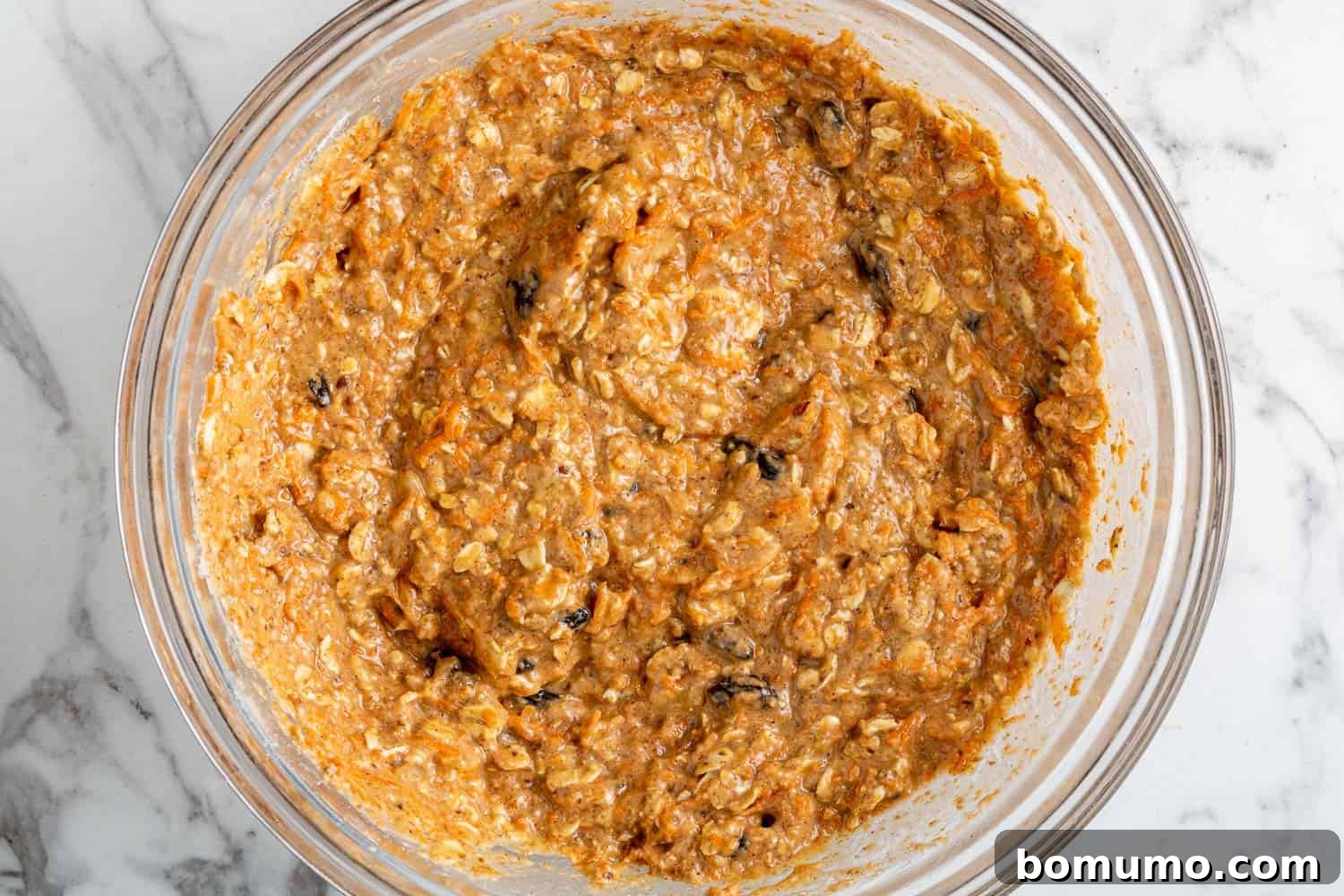 Muffin batter, with visible bits of carrot and raisin, in a clear glass mixing bowl, perfectly mixed.