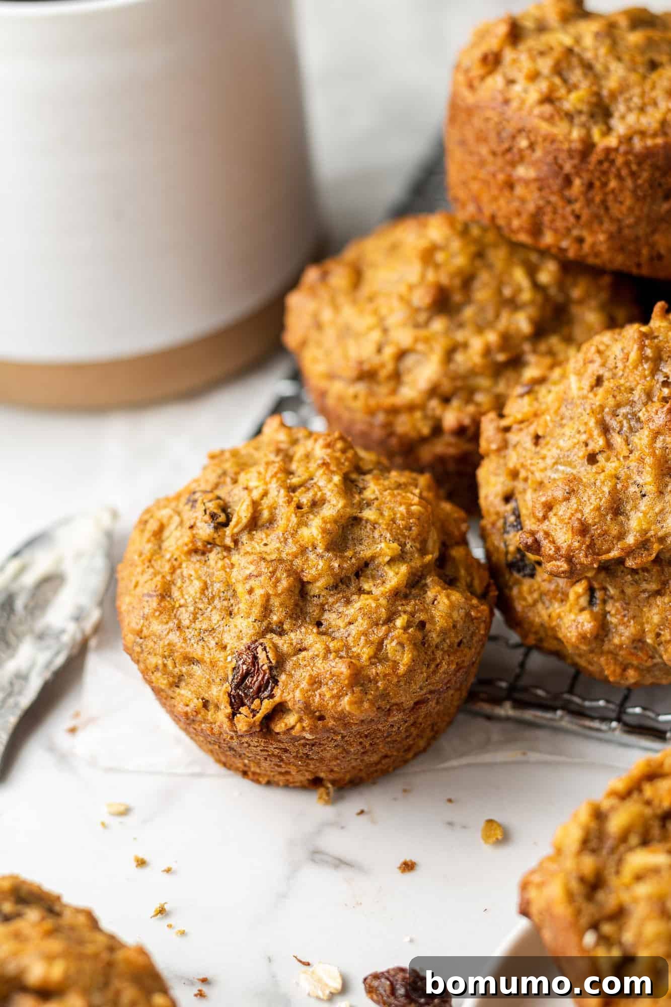 A rustic pile of healthy carrot raisin muffins arranged on a white surface, with a blurred coffee mug in the background, creating a cozy scene.