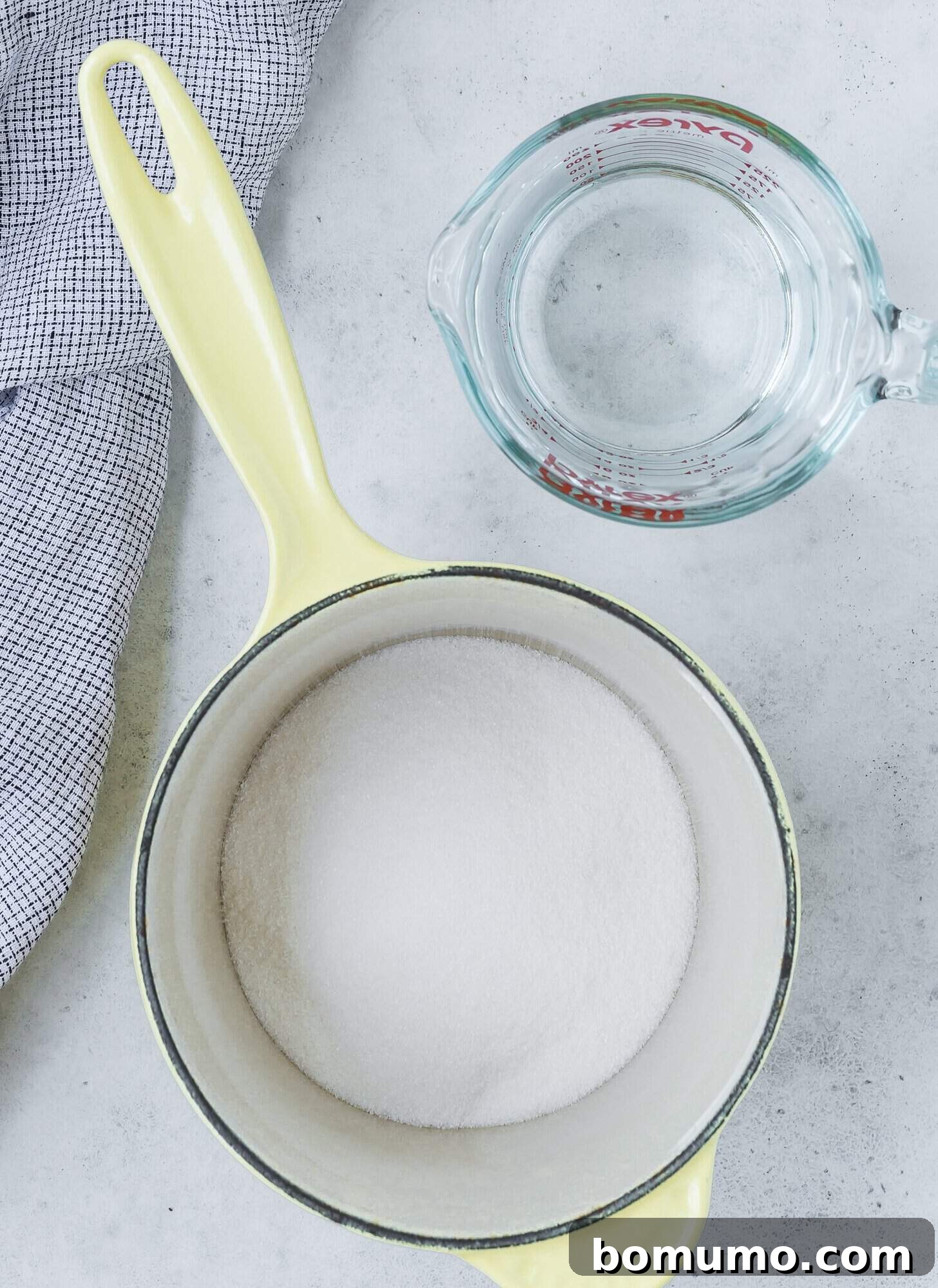 A pot with sugar inside next to a glass measuring cup of water on a countertop with a checkered cloth, illustrating the simple ingredients needed for simple syrup.