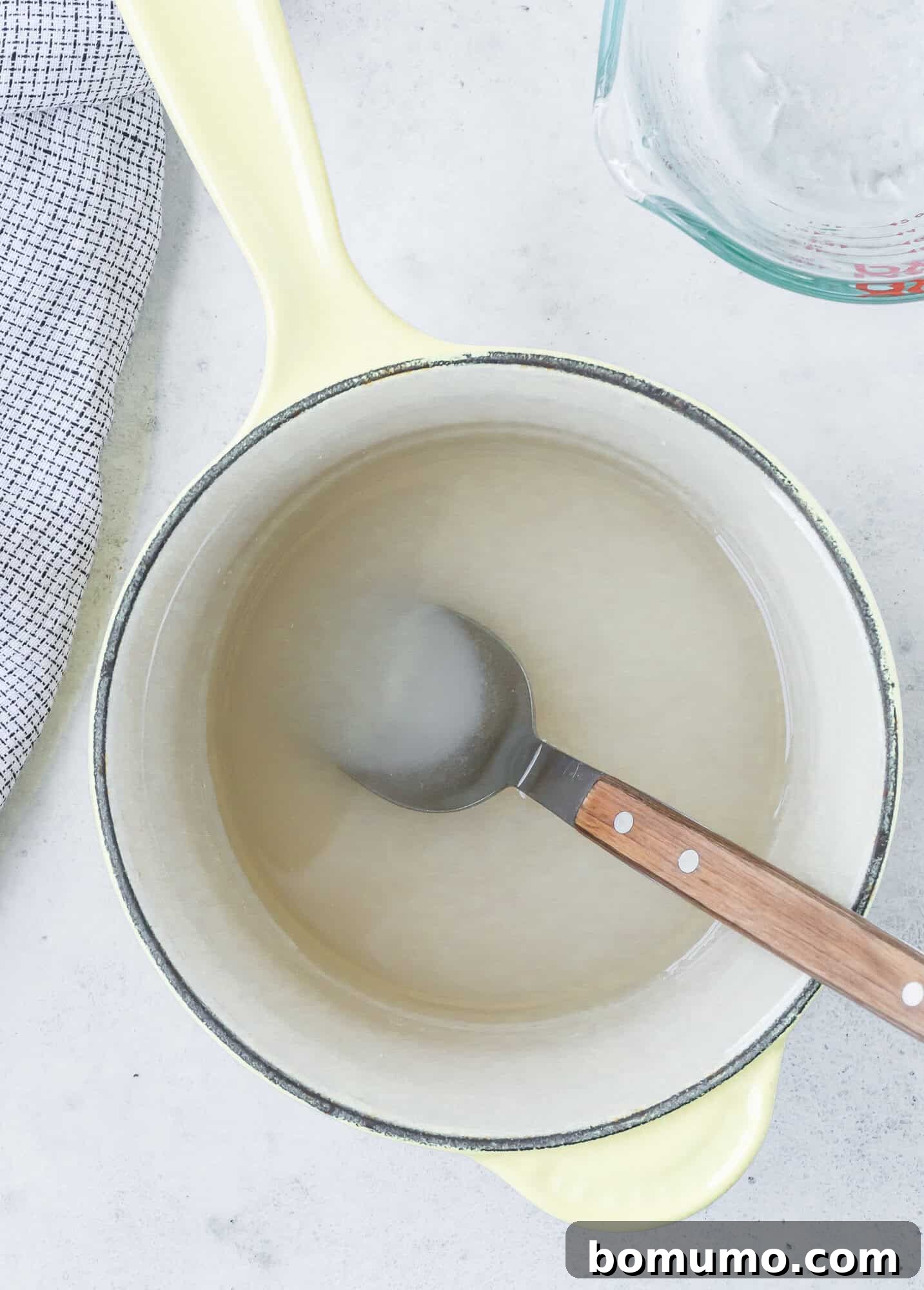 A spoon rests in a pot containing a translucent liquid, with a measuring cup nearby and a checkered cloth to the side, showcasing the dissolved sugar of simple syrup.