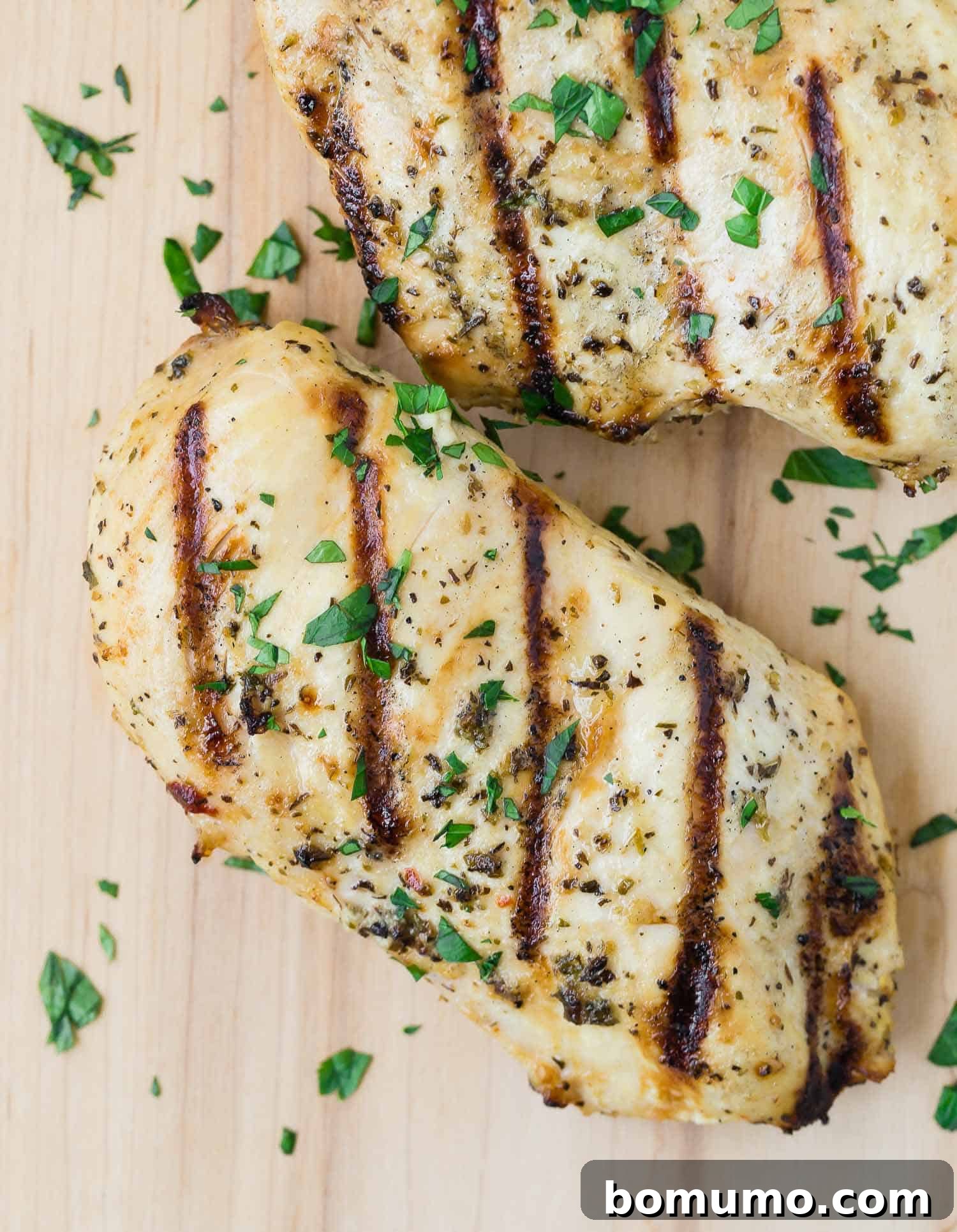Overhead view of grilled chicken breasts on a wooden cutting board, sprinkled with fresh parsley.