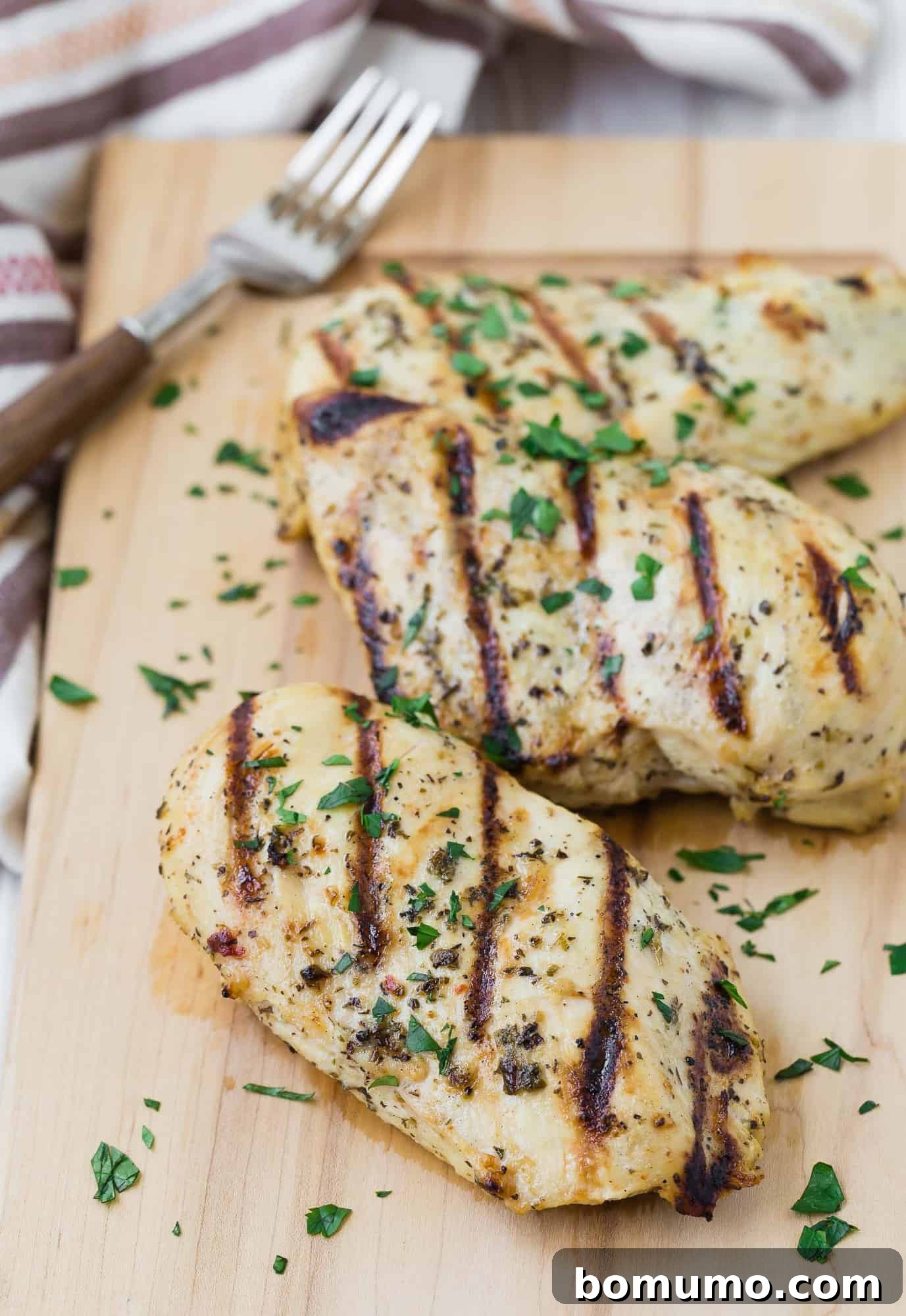 Three grilled boneless skinless chicken breasts on a wooden cutting board with a fork in the background.