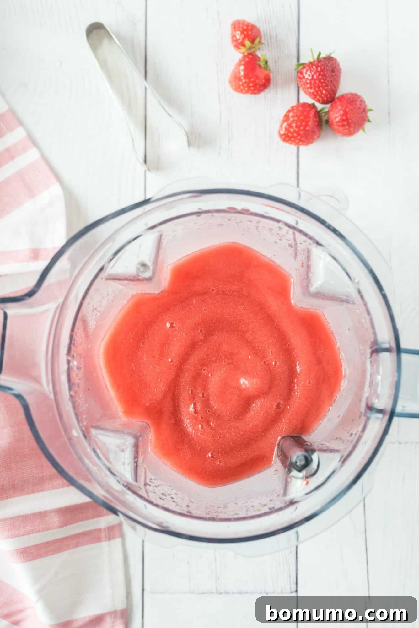 A close-up of a blender filled with freshly made, smooth Frosé, resting on a clean white wooden surface. Fresh whole strawberries and a pink-striped cloth add a touch of color and charm to the scene, highlighting the freshness of the ingredients.