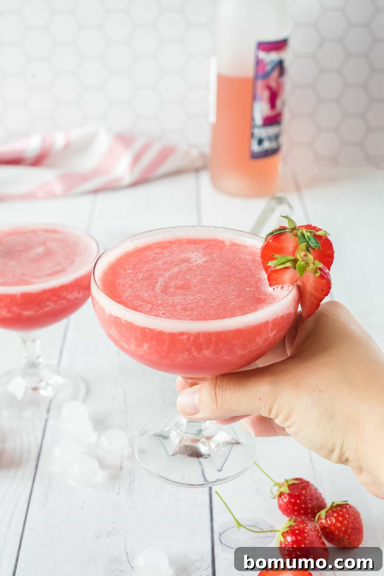 A hand gracefully holds a glass of the inviting pink Frosé cocktail, garnished with a vibrant fresh strawberry, highlighting its refreshing appeal and readiness to be enjoyed.