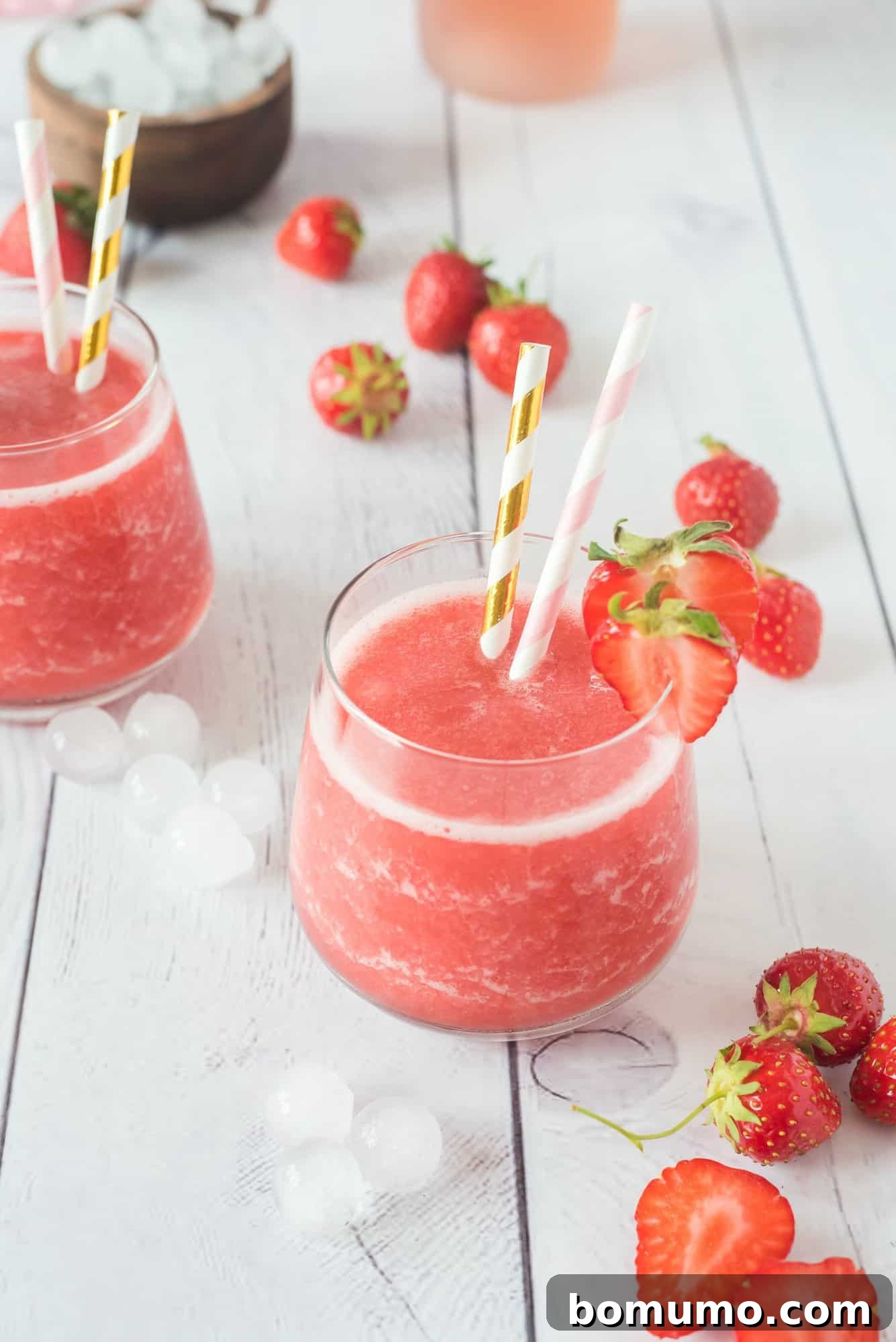 Two elegant glasses of strawberry Frosé, beautifully garnished with fresh strawberries and charming pink and white striped straws, are artfully arranged on a rustic white wooden table, ready for enjoyment.